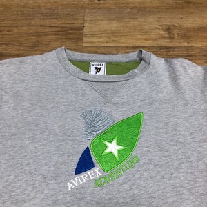 Puede incluir: Sudadera gris con un logotipo bordado en verde, azul y blanco que dice "Avirrex Adventure". El logotipo presenta una estrella y una ola estilizada. La sudadera tiene una etiqueta verde que dice "Avirrex" en el interior del cuello.