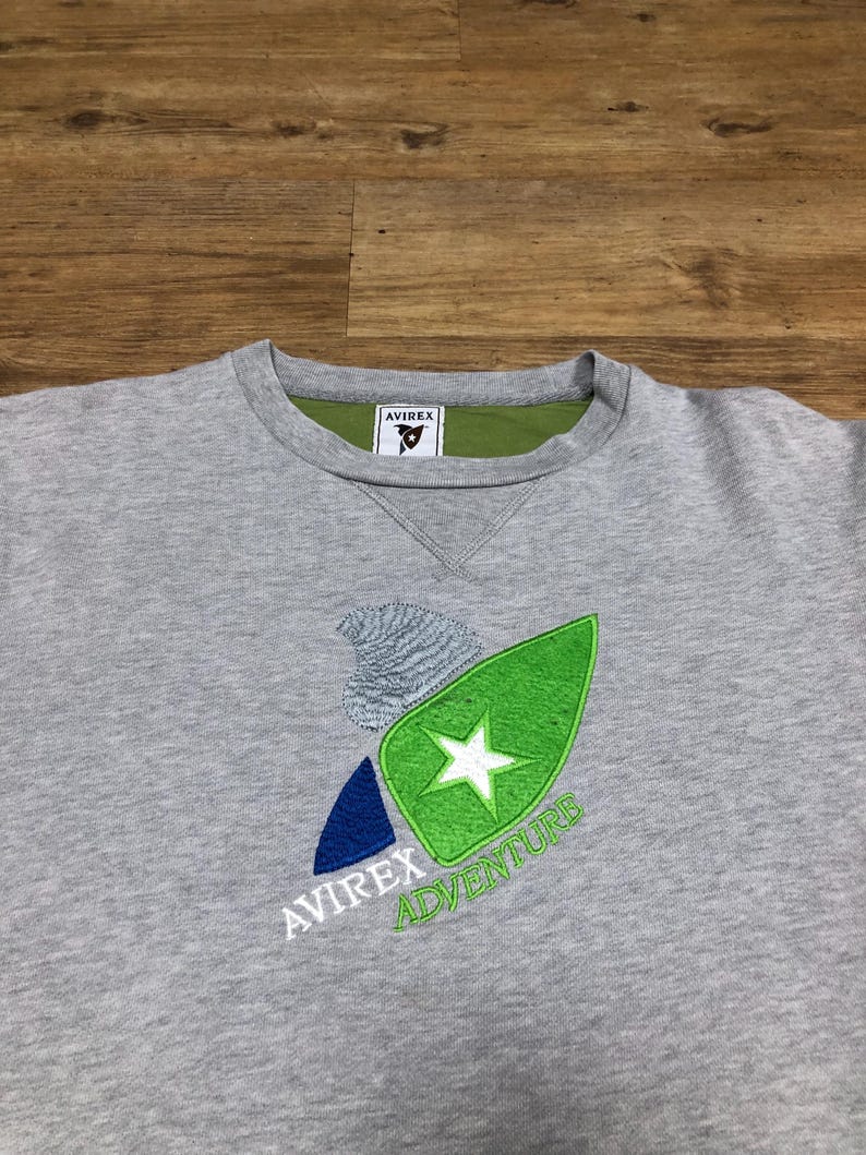 Puede incluir: Sudadera gris con un logotipo bordado en verde, azul y blanco que dice "Avirrex Adventure". El logotipo presenta una estrella y una ola estilizada. La sudadera tiene una etiqueta verde que dice "Avirrex" en el interior del cuello.