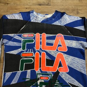 May include: A blue, black, and white sweatshirt with a large, orange "FILA" logo printed on the front. The logo is repeated twice, with the second "FILA" logo printed in green and red.