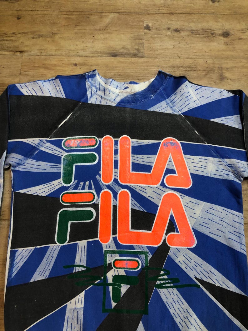 May include: A blue, black, and white sweatshirt with a large, orange "FILA" logo printed on the front. The logo is repeated twice, with the second "FILA" logo printed in green and red.