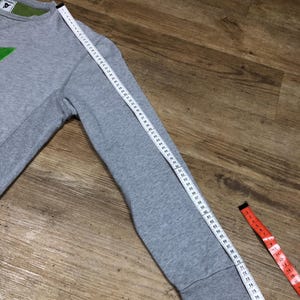 Puede incluir: Sudadera gris con un gr&aacute;fico verde en la parte delantera. La sudadera tiene un cuello redondo de canal&eacute; y mangas largas. La longitud de la manga se mide con una cinta m&eacute;trica.