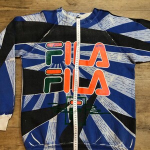 May include: A blue, black, and white sweatshirt with a large orange and green "FILA" logo. The sweatshirt has a geometric pattern and is made of a soft, comfortable fabric.