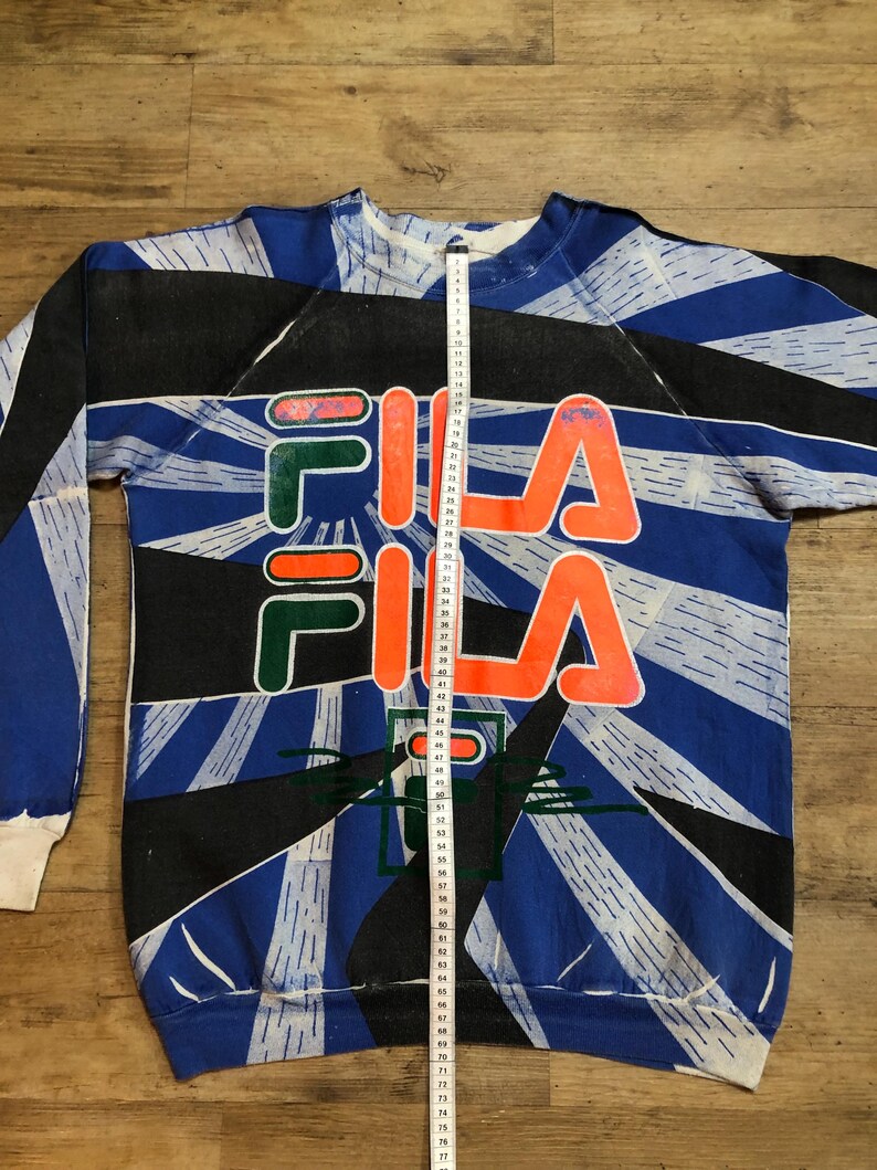 May include: A blue, black, and white sweatshirt with a large orange and green "FILA" logo. The sweatshirt has a geometric pattern and is made of a soft, comfortable fabric.