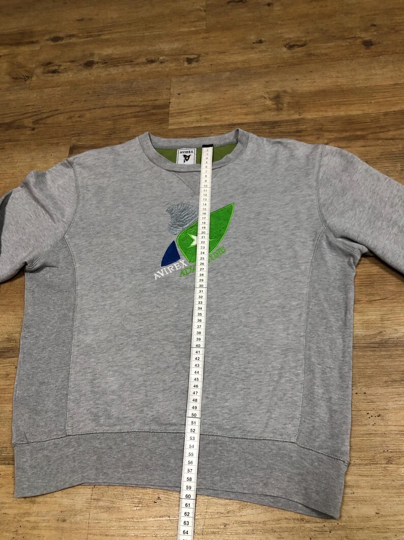 Puede incluir: Sudadera gris con un logotipo bordado en verde, azul y blanco que dice "AVIREX" y "Adventure".