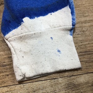 May include: Close-up of the cuff of a white long-sleeve shirt with blue paint splatters. The cuff is made of a ribbed fabric.