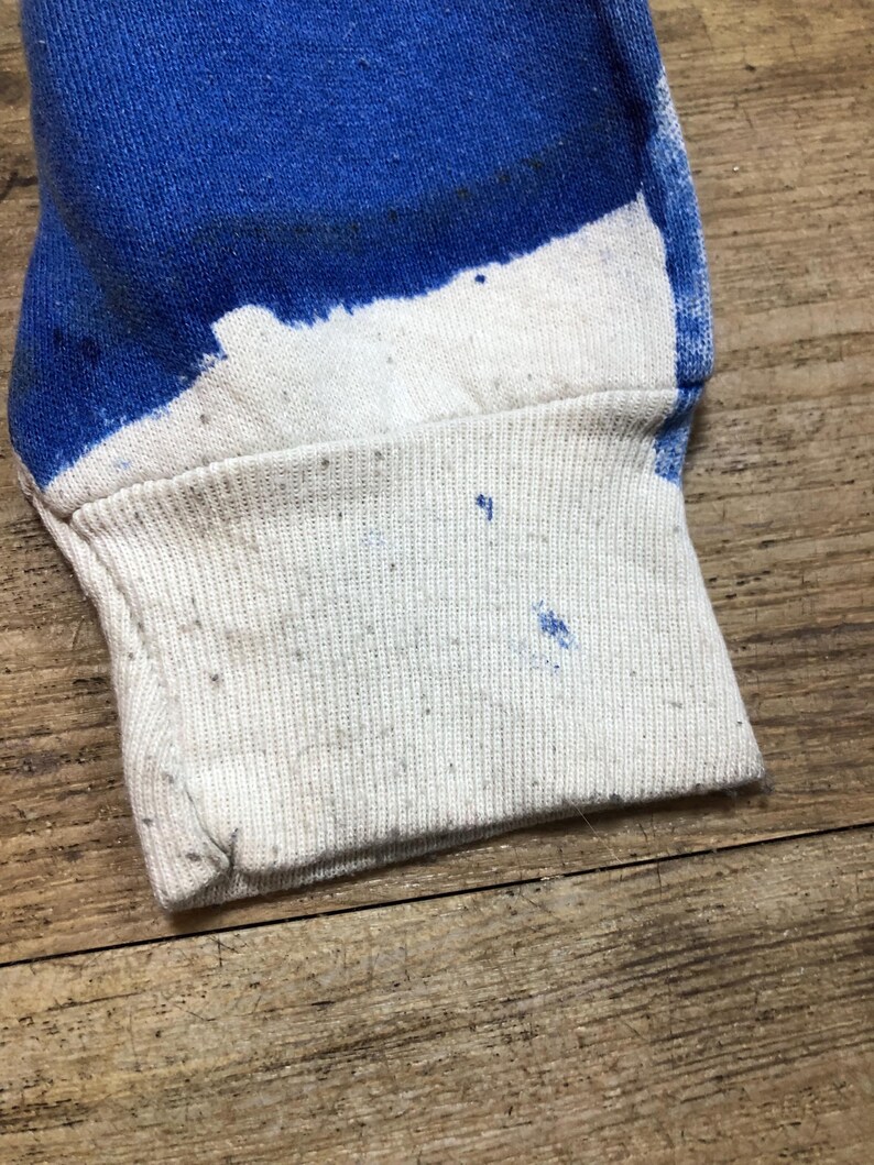 May include: Close-up of the cuff of a white long-sleeve shirt with blue paint splatters. The cuff is made of a ribbed fabric.