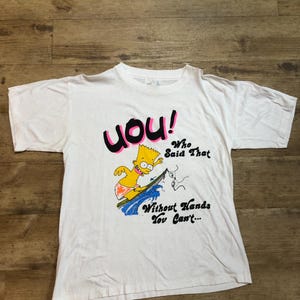 Könnte beinhalten: Weißes T-Shirt mit einem Cartoon-Bild eines gelben Charakters beim Surfen. Der Text auf dem Shirt lautet "YOU! Who Said That Without Hands You Can't..."