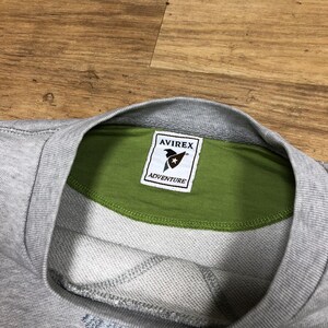 Puede incluir: Una sudadera gris con un cuello interior verde y un parche marr&oacute;n con el texto "AVIREX" y una estrella sobre el texto "ADVENTURE".