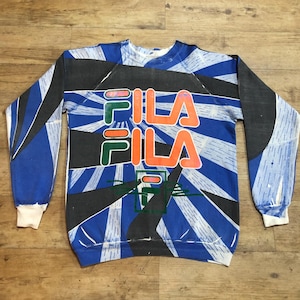 May include: A blue, black, green, and orange sweatshirt with a large "FILA" logo printed on the front. The sweatshirt has a vintage style and a faded look.