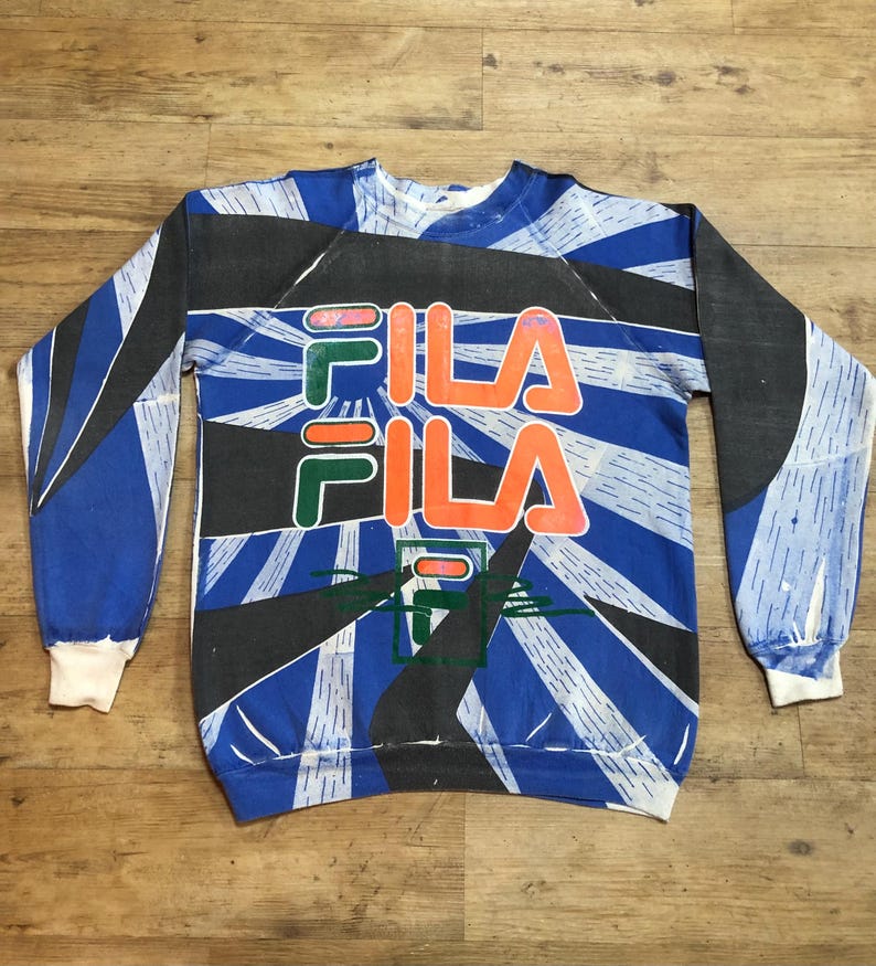 May include: A blue, black, green, and orange sweatshirt with a large "FILA" logo printed on the front. The sweatshirt has a vintage style and a faded look.