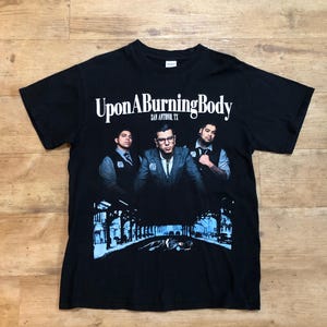 Könnte beinhalten: Schwarzes T-Shirt mit einem Band-Motiv, das drei Männer in Anzügen zeigt. Der Bandname ist "Upon A Burning Body" und der Text "San Antonio, TX" befindet sich unter dem Bandnamen.