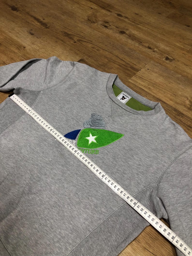 Puede incluir: Sudadera gris con un logotipo bordado en verde y azul que presenta una estrella y la palabra "AVIREX".