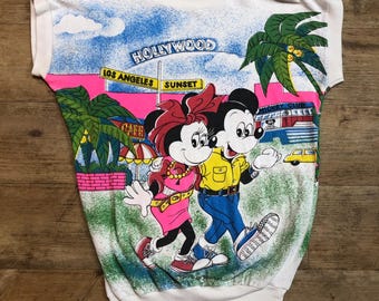 Könnte beinhalten: Weißes T-Shirt mit einem bunten Aufdruck von Mickey und Minnie Mouse, die eine Straße in Hollywood, Kalifornien, entlanglaufen. Der Aufdruck enthält Palmen, Gebäude und den Text "Hollywood", "Los Angeles" und "Sunset".