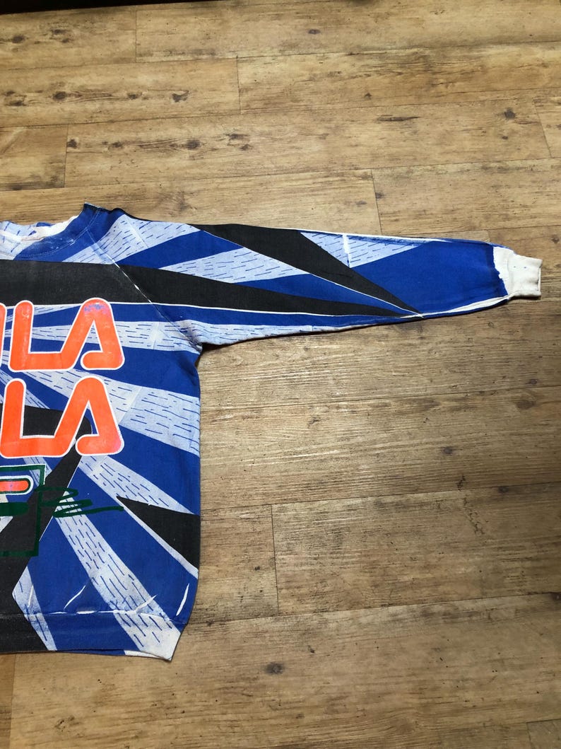 May include: A blue, black, and white long-sleeved sweatshirt with a graphic print of the word "FILA" in orange and white.