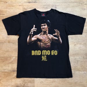 Könnte beinhalten: Schwarzes T-Shirt mit einem Grafikdruck von Bruce Lee in Kampfpose. Unter dem Bild ist der Text "Bad Mo Fo" und ein chinesisches Schriftzeichen gedruckt.