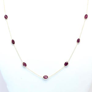Puede incluir: Un delicado collar de oro con piedras preciosas ovaladas de color rojo intenso, espaciadas uniformemente a lo largo de la cadena. El collar se muestra sobre un fondo blanco, resaltando el contraste.