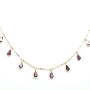 May include: A delicate gold necklace featuring multiple teardrop-shaped, deep purple gemstones. The gemstones are evenly spaced along the fine gold chain, creating a graceful and elegant design. The necklace is set against a clean white background.