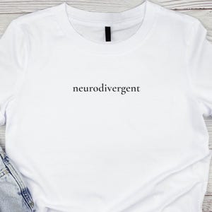 May include: A white t-shirt featuring the word "neurodivergent" in black, centrally printed. The shirt has a crew neck and short sleeves. The fabric appears soft, and the garment has a slightly creased appearance. The shirt is likely made of cotton.