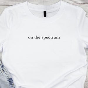 May include: White t-shirt featuring the text "on the spectrum" in black, centrally placed. The top has a round neck and short sleeves. A portion of blue jeans is visible at the bottom of the image.