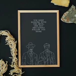 May include: Framed art print featuring a black background with white text: "YOU KEEP DANCING WITH THE DEVIL, ONE DAY HE'S GONNA FOLLOW YOU HOME." Below the text are two line drawings of men in suits and hats. The frame is light wood.