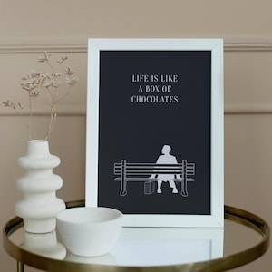 Op de afbeelding: Wit ingelijste kunstdruk met de quote "LIFE IS LIKE A BOX OF CHOCOLATES" op een donkere achtergrond. De print toont een silhouet van een persoon die met een koffer op een bank zit. Een witte vaas en kom staan op een gouden tafel.