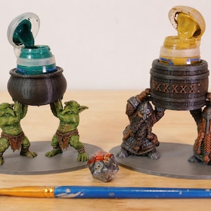 May include: Two miniature figurine sets holding paint pots: one with two green goblins supporting a black cauldron, the other with two armored dwarves holding a brown barrel. A multi-faceted red and silver die and a blue paintbrush lie in front.