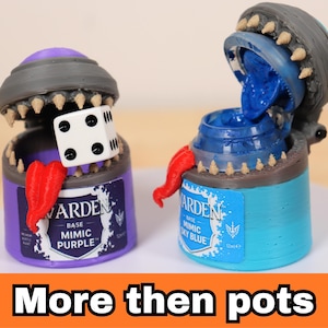 May include: Two cylindrical containers shaped like monsters with open lids, sharp teeth, and red tongues. The purple container is labeled WARDEN BASE MIMIC PURPLE and holds a white six-sided die. The blue container is labeled WARDEN BASE MIMIC SKY BLUE.