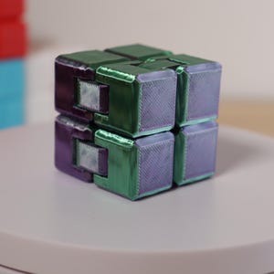 May include: A multi-colored infinity cube, featuring green and purple sections. The cube is on a white surface, with other cubes visible in the background. The cube is a fidget toy.