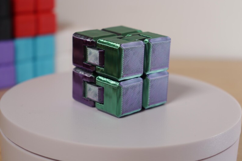 May include: A multi-colored infinity cube, featuring green and purple sections. The cube is on a white surface, with other cubes visible in the background. The cube is a fidget toy.