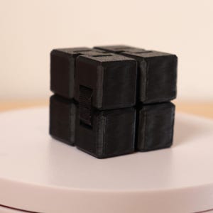 May include: A black fidget cube, composed of eight connected cubes, sits on a white surface. The cube is made of a matte material and has a textured appearance. The design allows for folding and unfolding.