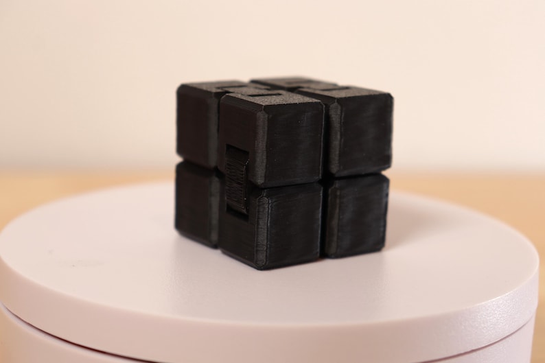 May include: A black fidget cube, composed of eight connected cubes, sits on a white surface. The cube is made of a matte material and has a textured appearance. The design allows for folding and unfolding.
