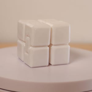 May include: A white, interlocking cube puzzle, composed of smaller square blocks. The puzzle is displayed on a light pink, circular platform. The background is a soft, neutral tone.