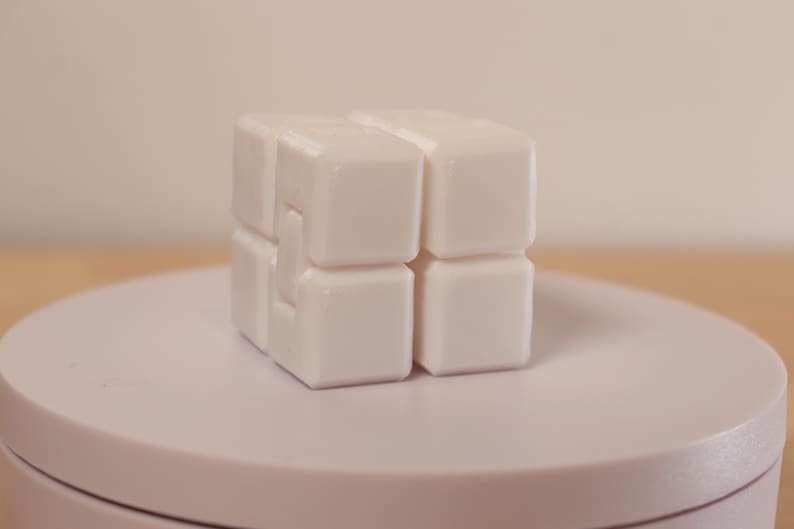 May include: A white, interlocking cube puzzle, composed of smaller square blocks. The puzzle is displayed on a light pink, circular platform. The background is a soft, neutral tone.