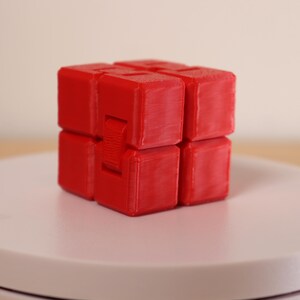 May include: A red infinity cube fidget toy. The cube is composed of eight connected blocks, each with a square shape. The toy is designed to be flipped and folded, providing a tactile and engaging experience. The cube is sitting on a white surface.