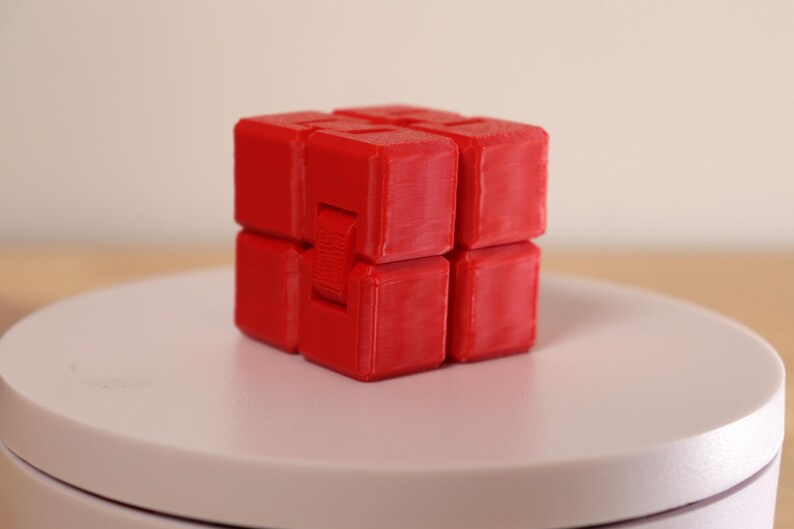 May include: A red infinity cube fidget toy. The cube is composed of eight connected blocks, each with a square shape. The toy is designed to be flipped and folded, providing a tactile and engaging experience. The cube is sitting on a white surface.