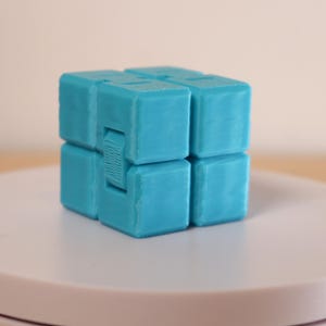 May include: A turquoise infinity cube fidget toy. The cube is made of several smaller cubes connected together, allowing for movement and manipulation. The toy is designed to relieve stress and improve focus. It is sitting on a white surface.