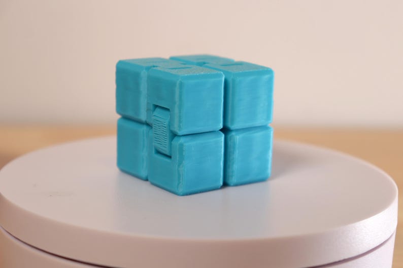 May include: A turquoise infinity cube fidget toy. The cube is made of several smaller cubes connected together, allowing for movement and manipulation. The toy is designed to relieve stress and improve focus. It is sitting on a white surface.