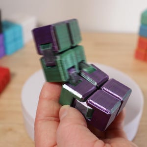 May include: A multi-colored fidget cube in shades of purple and green is held in a person's hand. Other colorful cubes are in the background, including red, yellow, black, blue, and orange. The cube is designed to be manipulated.