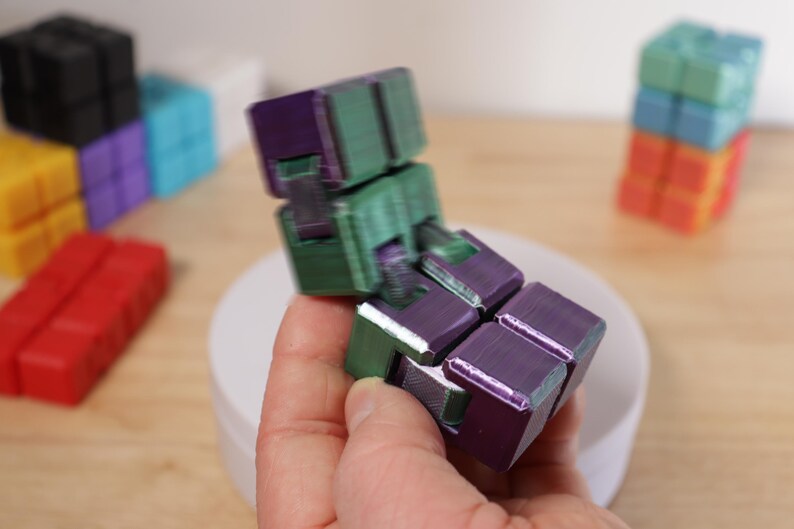 May include: A multi-colored fidget cube in shades of purple and green is held in a person's hand. Other colorful cubes are in the background, including red, yellow, black, blue, and orange. The cube is designed to be manipulated.