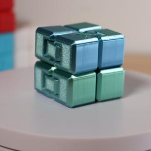 May include: A two-tone blue and green infinity cube, a fidget toy, is displayed on a white surface. The cube is made of several connected blocks. Other colorful cubes are visible in the background.