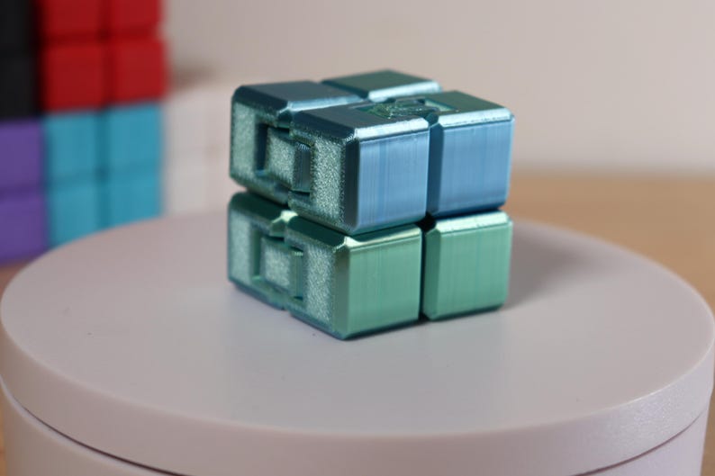 May include: A two-tone blue and green infinity cube, a fidget toy, is displayed on a white surface. The cube is made of several connected blocks. Other colorful cubes are visible in the background.