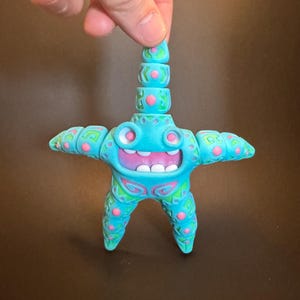 May include: A turquoise blue starfish figurine with pink and green details. The starfish has a smiling face with white teeth and pink cheeks. The figurine is standing on a brown surface.