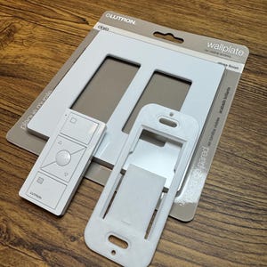 Puede incluir: Una placa de pared blanca Lutron con acabado brillante, un mando a distancia y un soporte de montaje. La placa de pared tiene dos aberturas y el texto "wallplate" y "no visible screws". El mando a distancia tiene varios botones y el logotipo de Lutron.