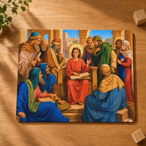 Può includere: Un puzzle rettangolare che raffigura una scena di una giovane persona con una tunica rossa seduta su un sedile di pietra, intenta a leggere un libro aperto, circondata da un gruppo di persone con tuniche di vari colori, tra cui blu, verde e marrone.