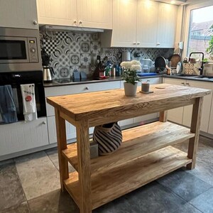 May include: A rectangular wooden kitchen island featuring a thick natural wood grain top and two lower open shelves. The island is constructed from light brown timber with visible wood grain patterns and stands on four sturdy legs.