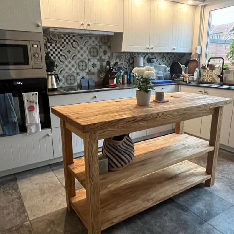 May include: A rectangular wooden kitchen island featuring a thick natural wood grain top and two lower open shelves. The island is constructed from light brown timber with visible wood grain patterns and stands on four sturdy legs.