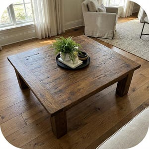 May include: A square coffee table crafted from dark brown wood with a visible grain and textured surface. Centered on the table is a black circular tray holding a small white pot with a green fern, a white rectangular book, and a small bowl.