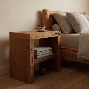 May include: A rustic wooden nightstand with a lower shelf and a thick top. The nightstand is made of light brown wood and has visible wood grain. A stack of folded gray blankets sits on the shelf. A bed with gray bedding is in the background.