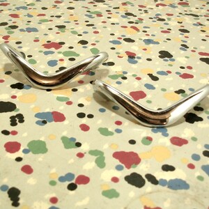 May include: Two shiny, silver, boomerang-shaped metal pieces on a speckled, light gray surface with red, black, blue, and yellow spots.