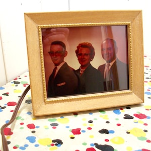 May include: A framed photograph of three people. The photo is in a light wood frame with a gold trim. The people in the photo are smiling and appear to be a family.